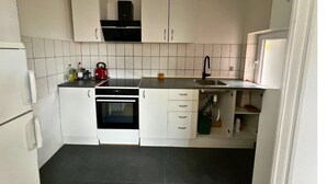 A compact kitchen with white cabinets, a black countertop, a built-in oven, and a sink. - Cozy Holeby Pet-friendly Villa - Puttgarden/fermen/lalandia/knuthenborg (Holeby)