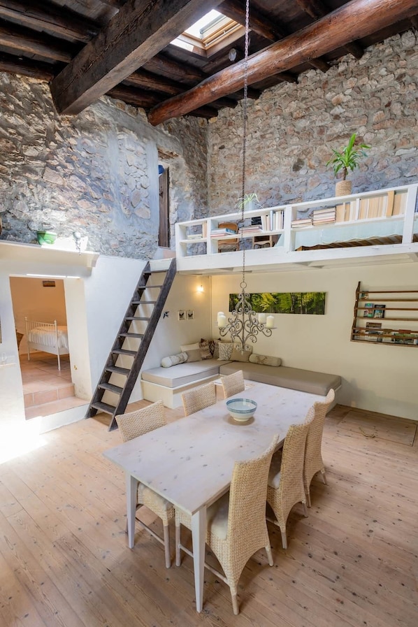 A rustic interior with exposed stone walls, a wooden staircase, a dining area with wicker furniture, and a loft with a plant.