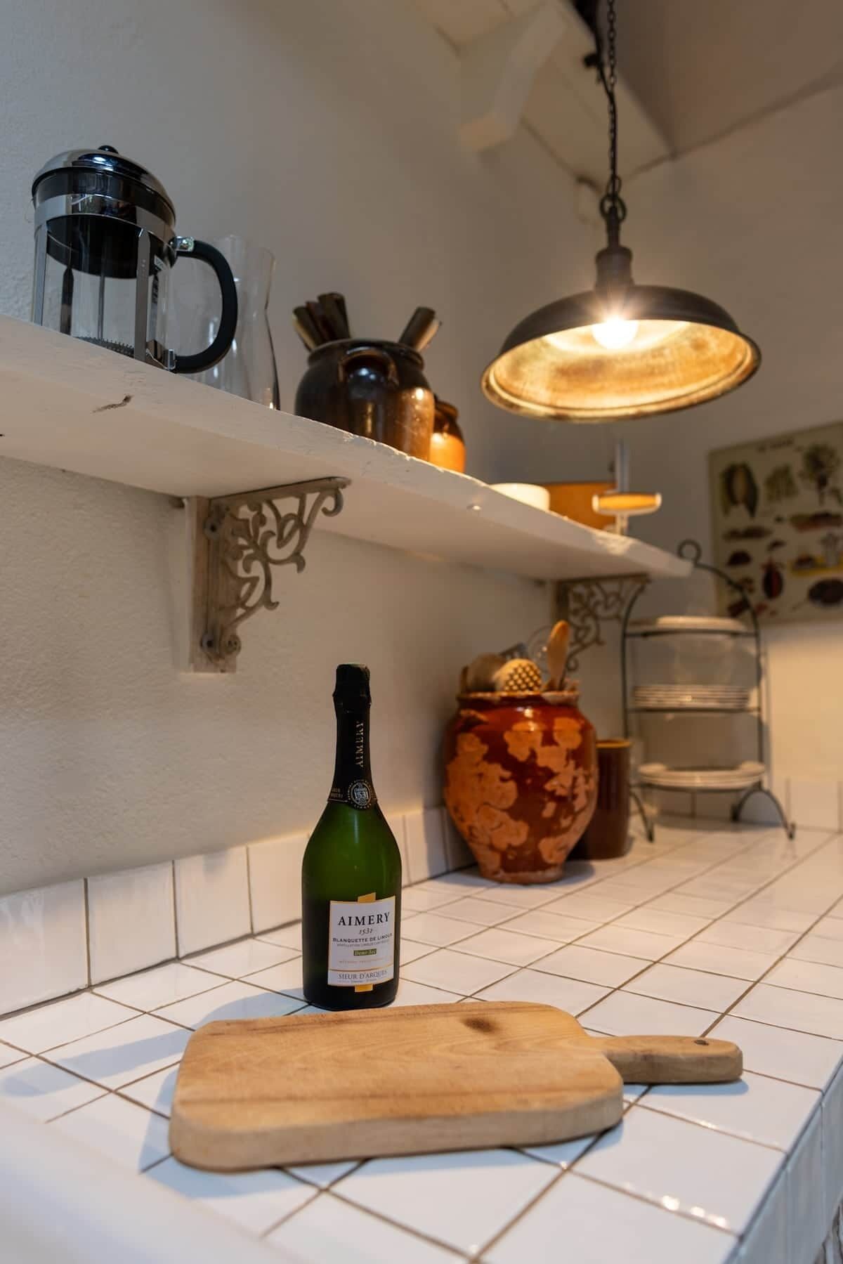 A kitchen with a wine bottle, a cutting board, a hanging light, and a shelf with kitchen items.