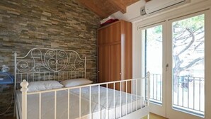 Un dormitorio con estructura de cama de metal blanco, pared de piedra, armario de madera y puerta corredera de vidrio que da a un espacio exterior.