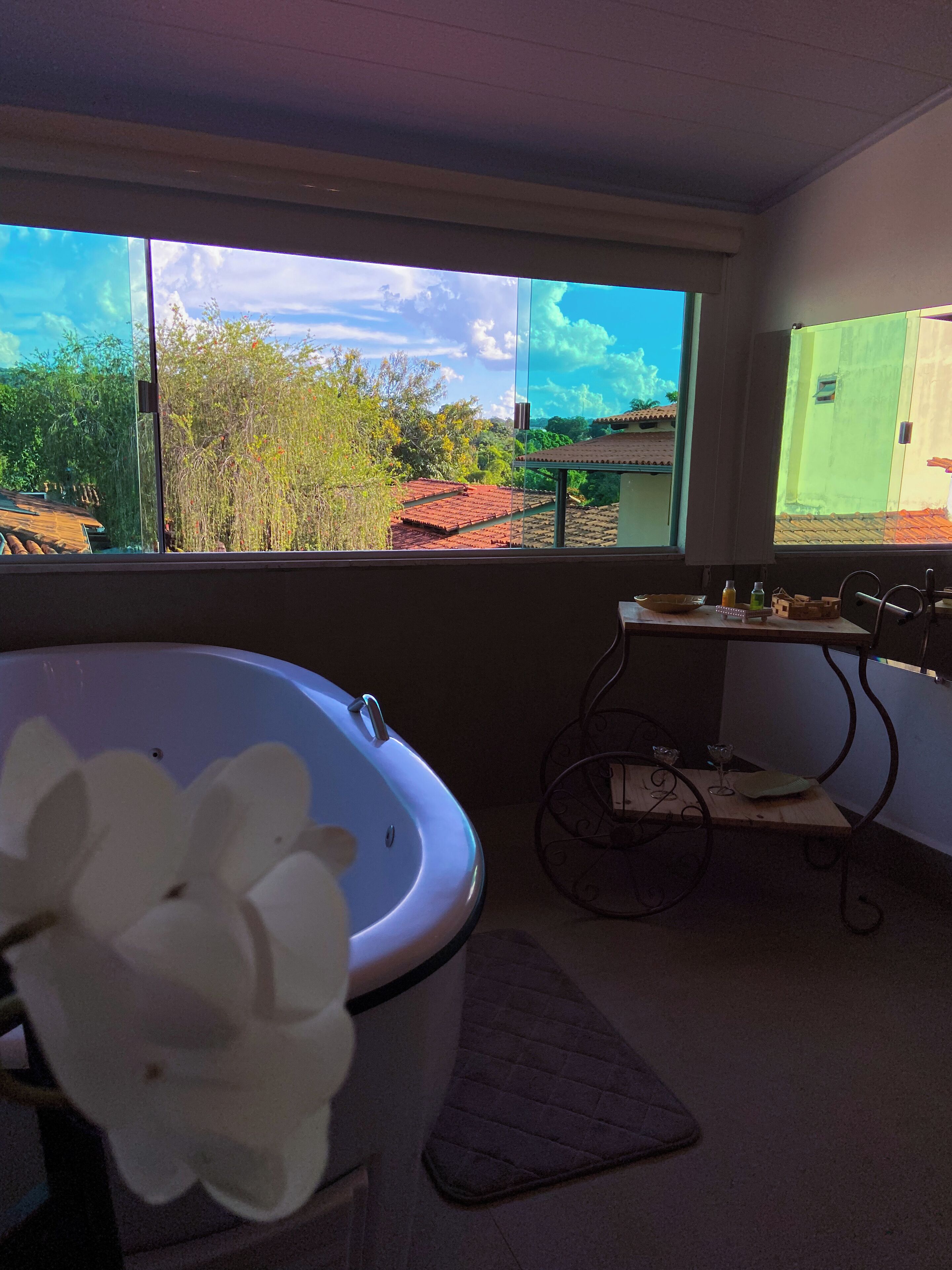 A bathroom with a bathtub, a window with a view of trees and buildings, and a small table with a glass and a bottle.