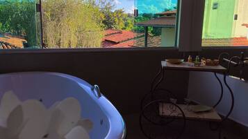 A bathroom with a bathtub, a window with a view of trees and buildings, and a small table with a glass and a bottle.