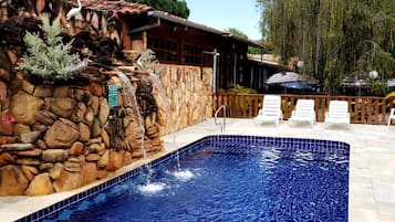 A swimming pool with a stone wall and a waterfall feature, surrounded by lounge chairs and a patio area.