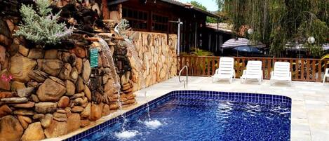 Una piscina con pared de piedra y cascada, rodeada de sillas de descanso y una zona de patio.