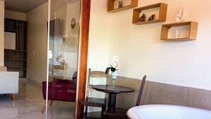 A modern room with a glass door leading to a balcony, a round table with chairs, a white bathtub, and wooden shelves with decorative items.