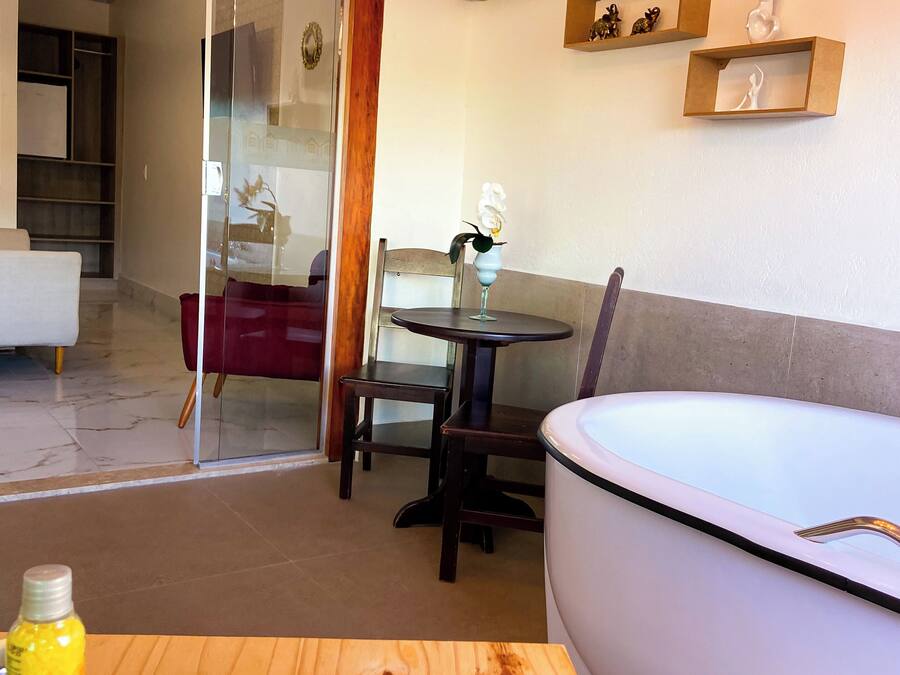 Una habitación moderna con puerta de cristal que da a un balcón, una mesa redonda con sillas, una bañera blanca y estantes de madera con objetos decorativos.