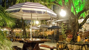 An outdoor seating area with wooden tables and benches, striped umbrellas, and a stone wall with plants.