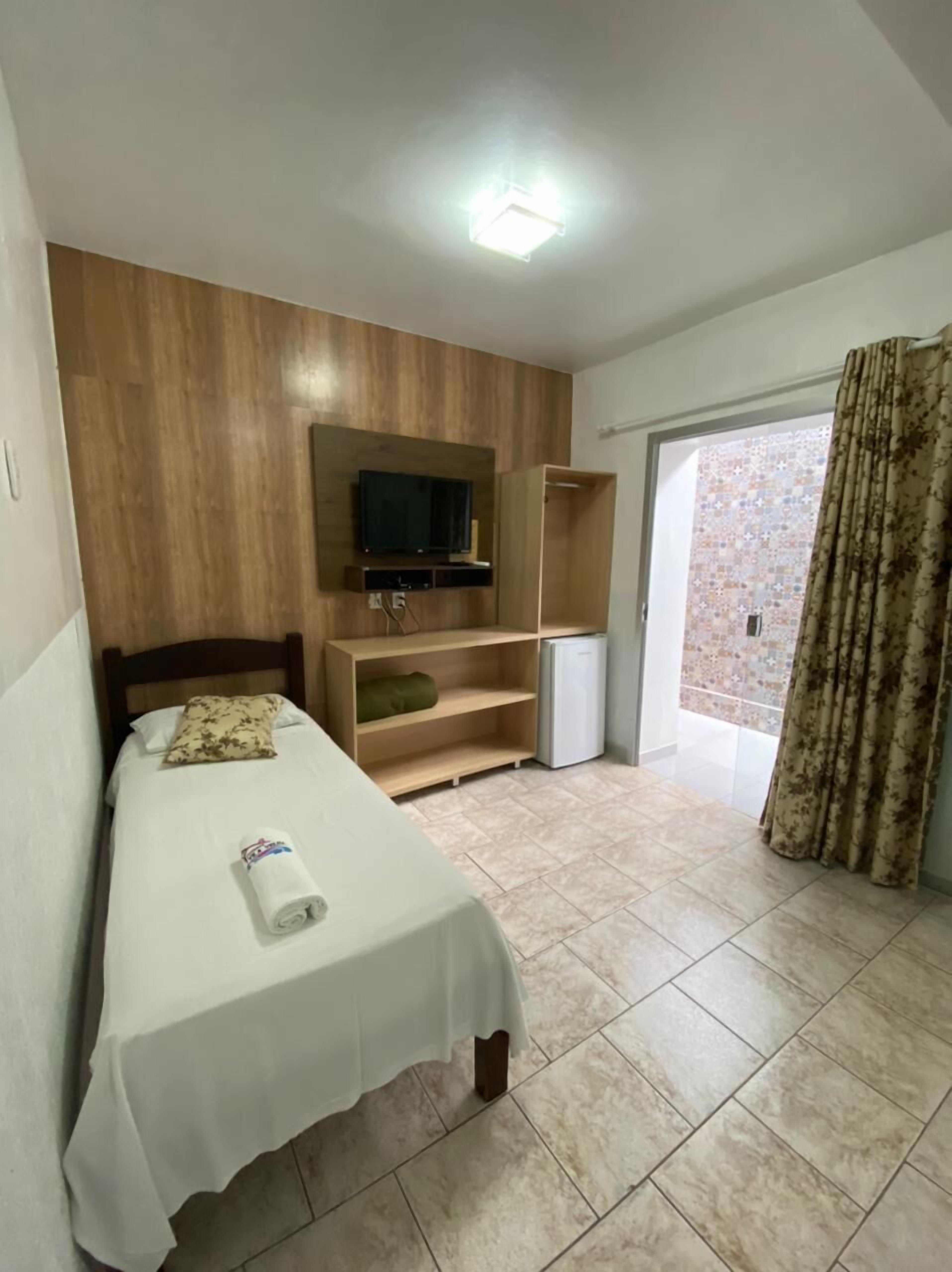 A hotel room with a bed, a TV mounted on a wooden panel, a small refrigerator, and a sliding door leading to a balcony.