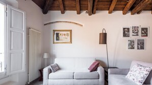A living room with a white sofa, a grey rug, and a wooden ceiling. There is a framed picture on the wall and a lamp on a side table. - Appartamento San Martino by Wonderful Italy (Bologna)