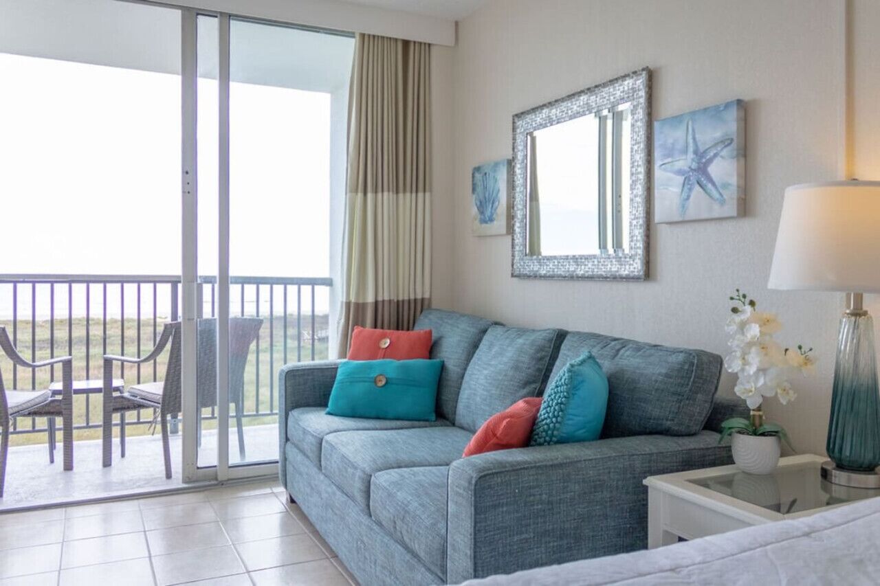 A room with a grey sofa, teal and red cushions, a white lamp, and a framed mirror on the wall.