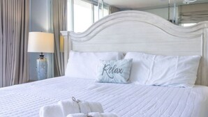 A neatly made bed with white linens and two pillows, one with the word "Relax" on it. There are two folded towels on the bed. - 911 Beachfront Heated Pool Gulf View 2 Bed 2 Bath (Galveston)