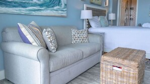 Une chambre avec un lit, un canapé gris agrémenté de coussins à motifs, un pouf en osier et un grand tableau représentant une vague océanique accroché au mur.
