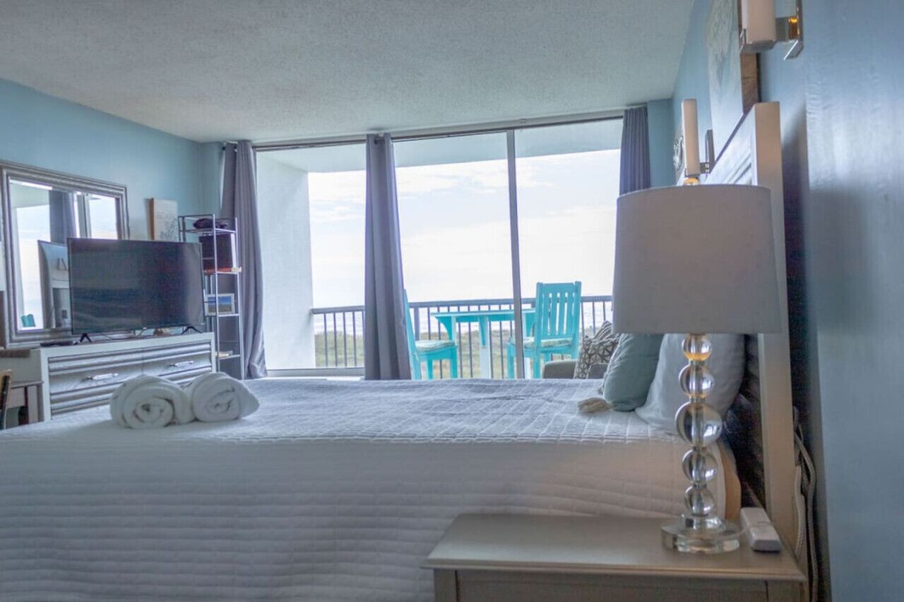 A hotel room with a large bed, a TV, a desk, and a balcony with chairs.