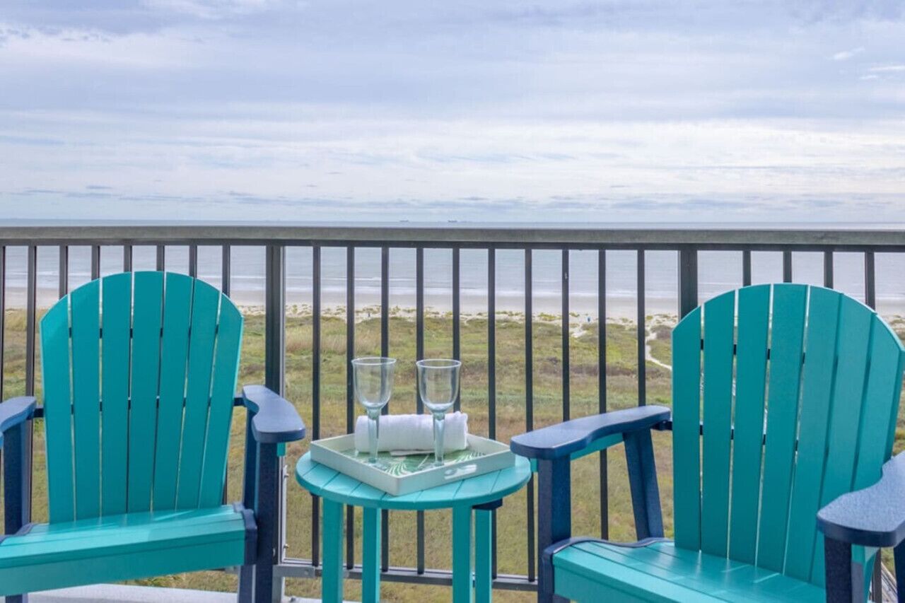 Zwei grüne Adirondack-Stühle mit einem kleinen Tisch und zwei Gläsern auf einem Balkon mit Blick auf einen Strand.