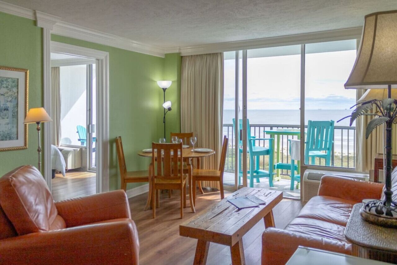 Une pièce avec une table et des chaises en bois, un canapé et une vue sur l’océan par de grandes fenêtres.