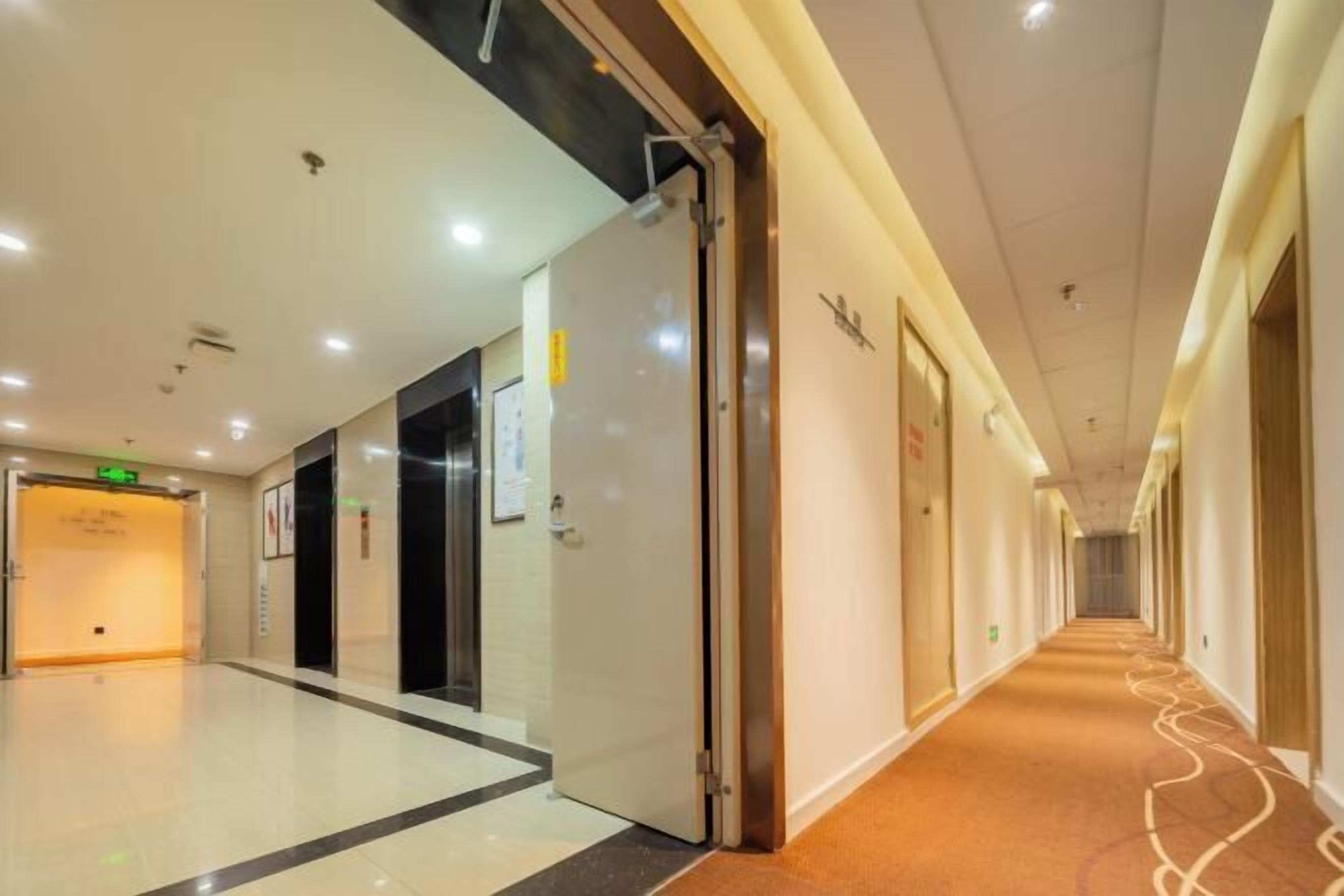 A hotel corridor with a carpeted floor, a glass elevator, and doors leading to rooms.