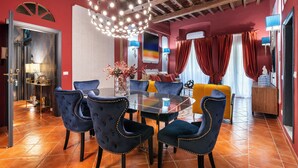 A dining area with a glass table surrounded by velvet chairs, a chandelier, and a red and yellow sofa in the background.