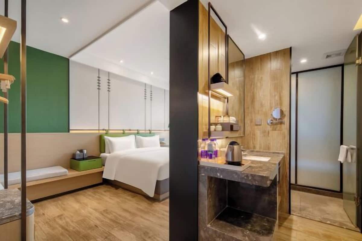 A modern hotel room with a bed, bedside table, green storage unit, and a bathroom with a sink, mirror, and shower.