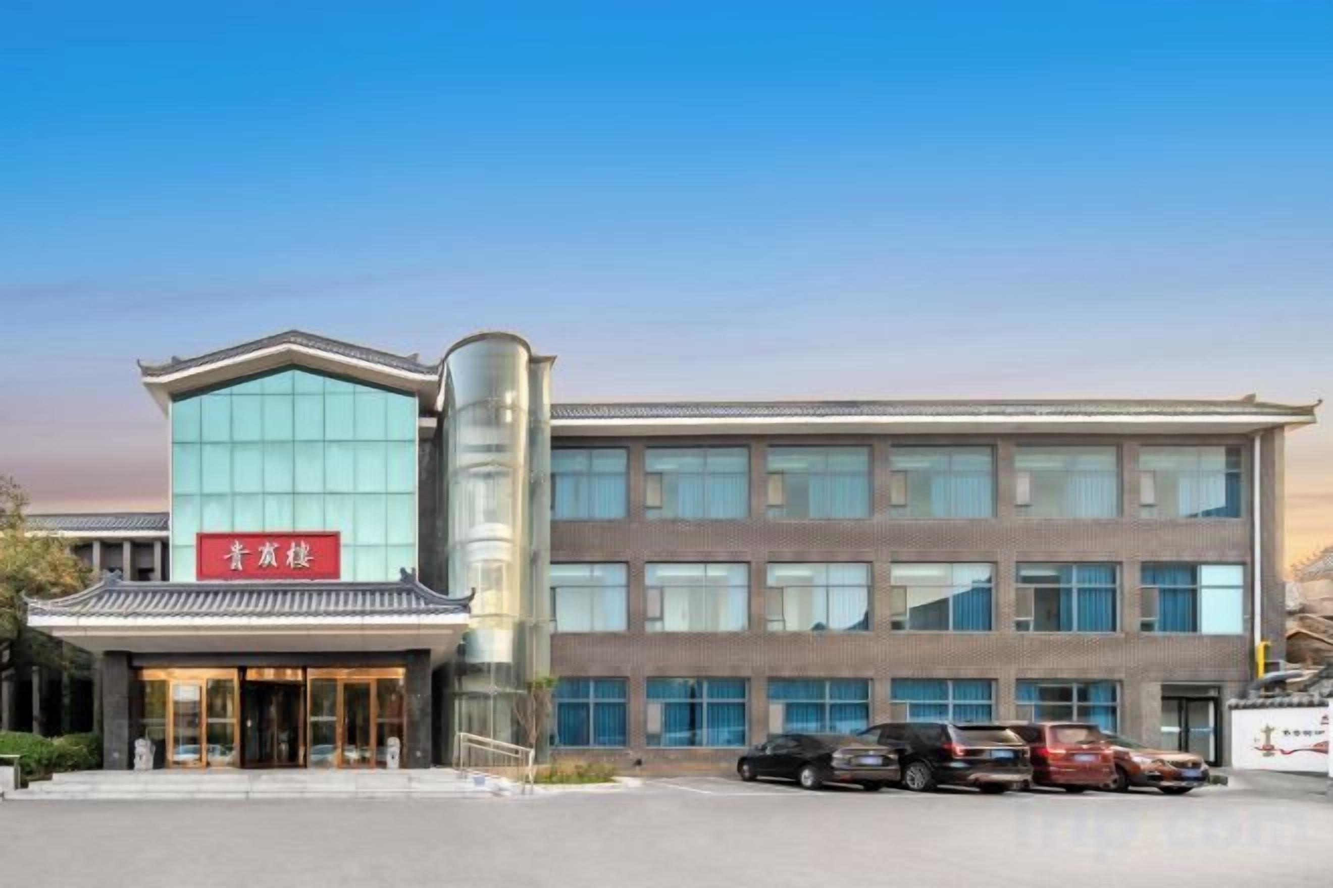 Un hotel de estilo arquitectónico tradicional, con una entrada destacada que tiene un letrero rojo, fachada de vidrio moderna y un estacionamiento con varios autos.