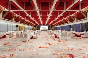 Un salón amplio con filas de mesas y sillas, un escenario al frente y piso alfombrado.