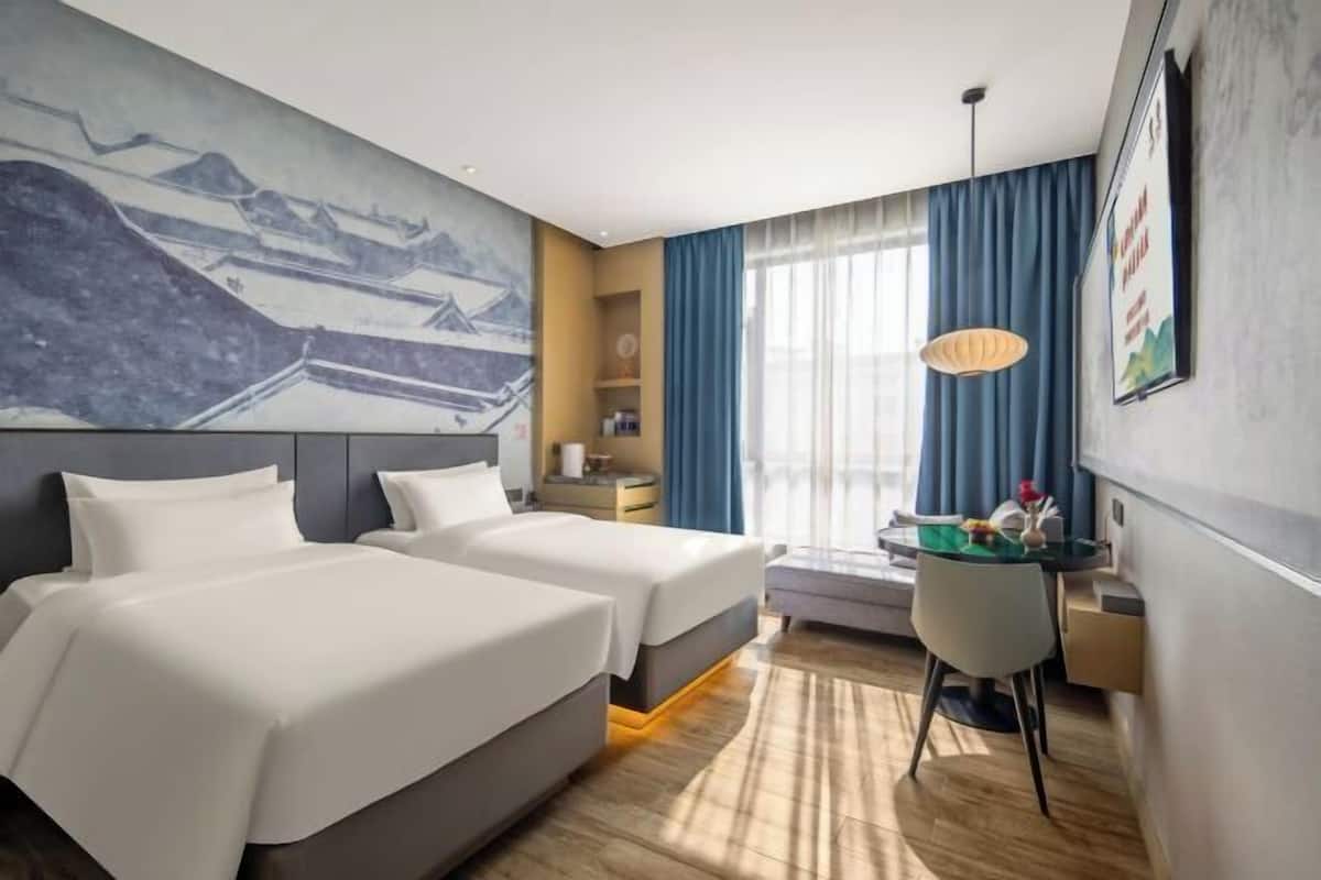 A hotel room with two beds, a desk, a chair, a large window with curtains, a painting on the wall, and a hanging light fixture.