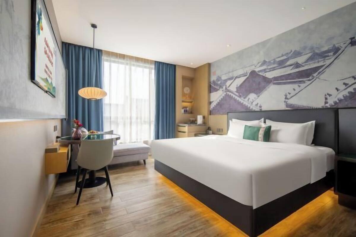 A hotel room with a large bed, a desk, a chair, a desk lamp, a window with curtains, and a wall painting.