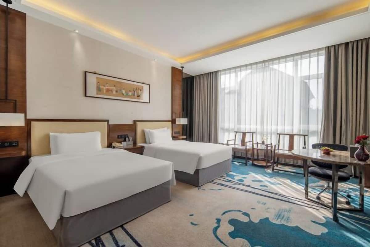 A hotel room with two beds, a desk, a chair, a dining table, and a large window with curtains.