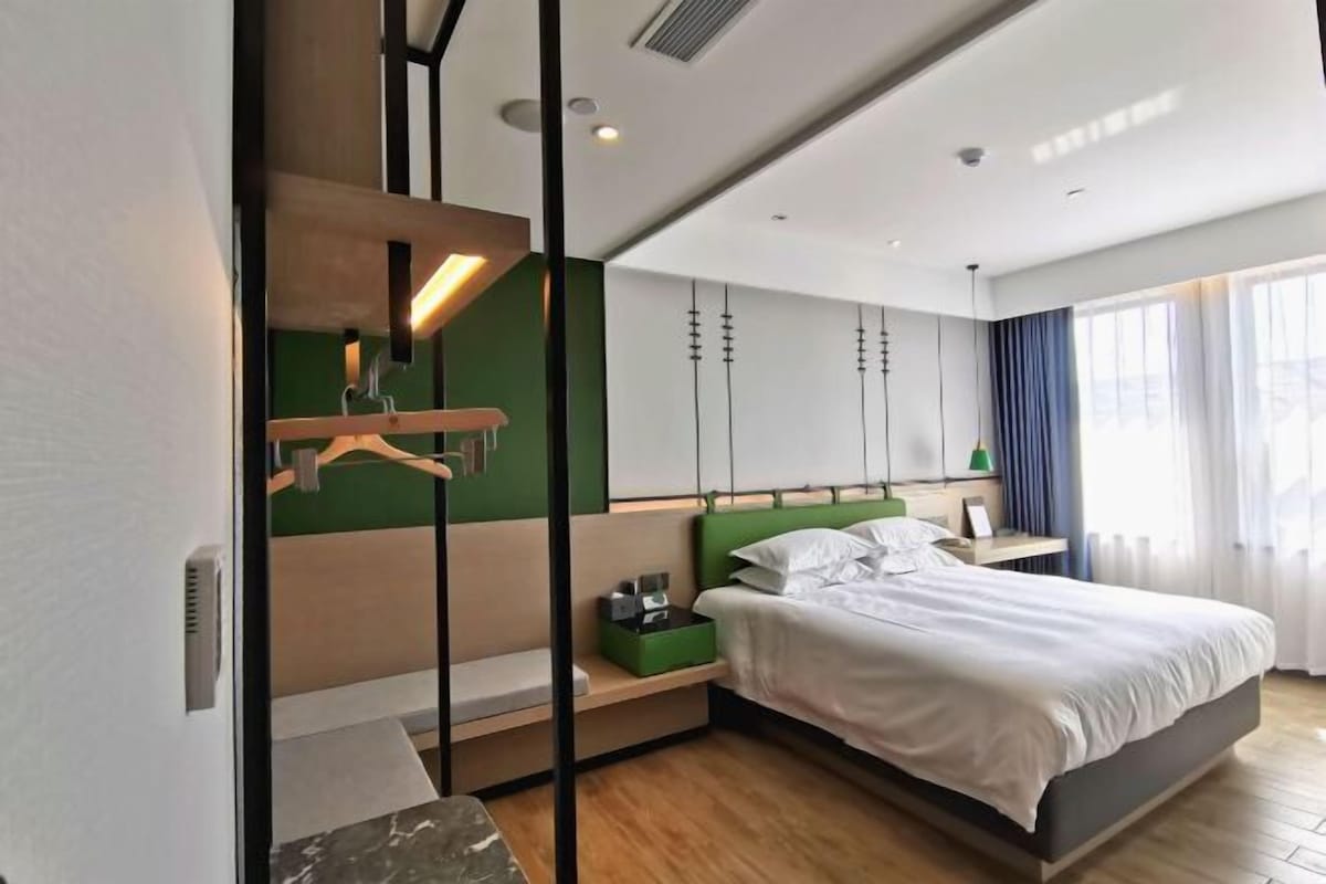 A modern hotel room with a bed, bedside tables, a green accent wall, and a large window with curtains.