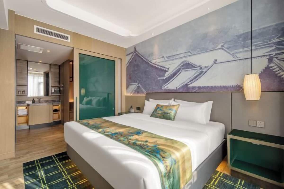 A hotel room with a bed, a painting of traditional architecture, a desk, and a green chair.