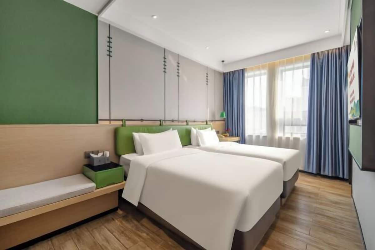 A hotel room with two beds, a bench, a green wall, and a large window with curtains.