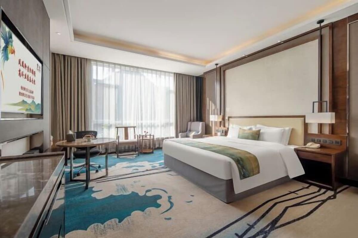 A hotel room with a large bed, a desk with a chair, a TV, and a large window with curtains.