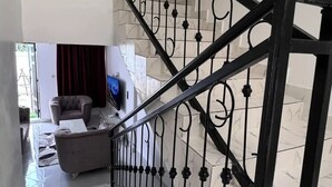 Una escalera con barandilla de metal negro, suelo de mármol y sala de estar con sofá y televisor.