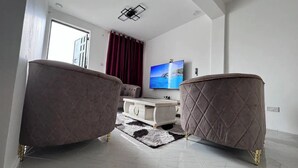 Un salón moderno con un televisor de pantalla plana grande, dos sillones, una mesa de centro blanca y una cortina roja.