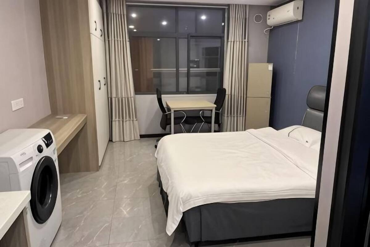 A bedroom with a bed, a desk, a chair, a washing machine, and a window with curtains.