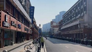 A street with buildings on either side, a clear sky, and a few vehicles.