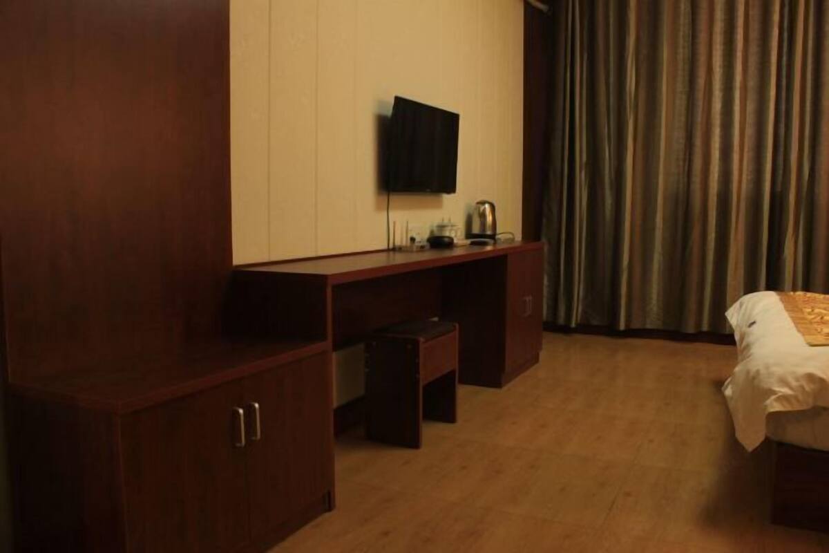 A hotel room with a wooden desk, a TV mounted on the wall, a bed with a white bedspread, and a window with curtains.