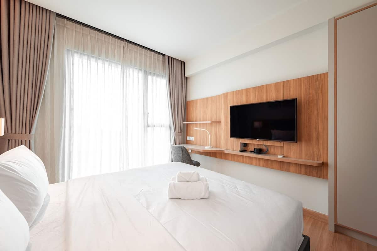 A hotel room with a large bed, a flat-screen TV mounted on a wooden panel, a desk with a lamp, and a chair.