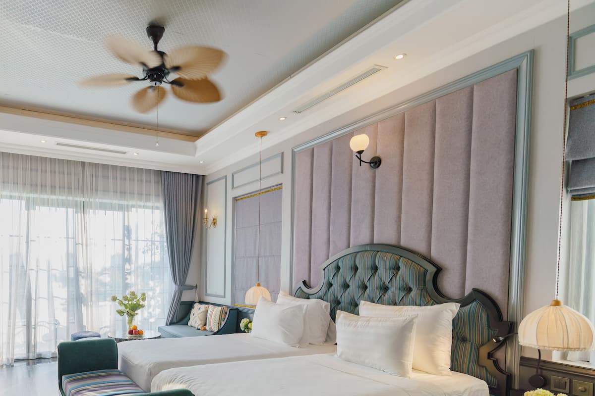 A hotel room with two beds, a ceiling fan, a large window with curtains, and a wall-mounted lamp.