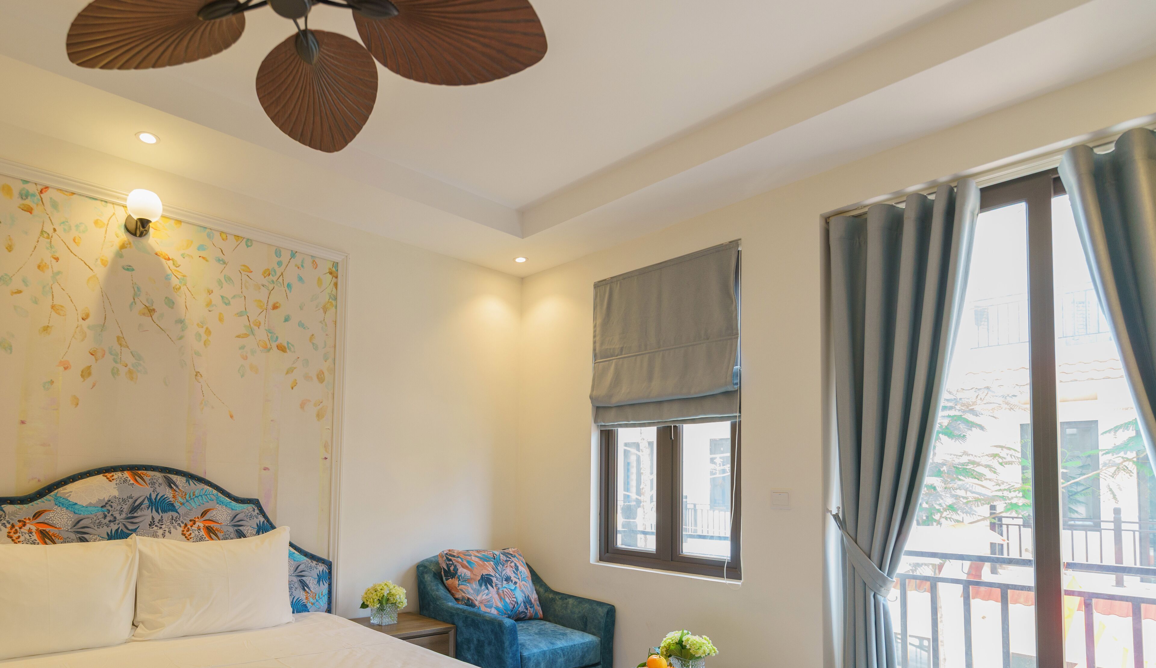 Un dormitorio con una cama, una silla, un ventanal con cortinas, una pared con diseño de hojas y un ventilador de techo.