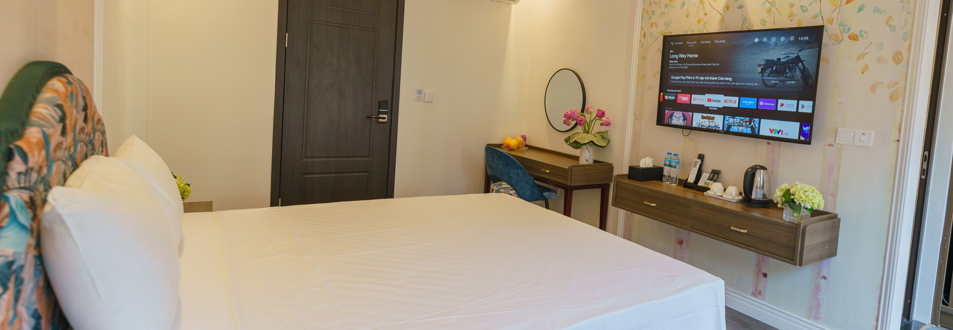 A hotel room with a bed, a desk with a computer monitor displaying various apps, a mirror, and a small table with a vase of flowers. - Lotus Hotel - Vuon vua (Tu Vu)