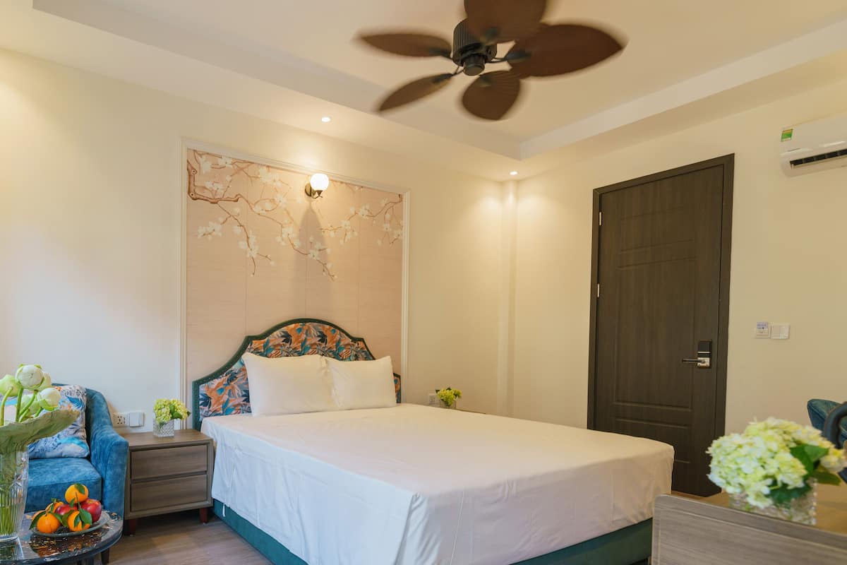 A bedroom with a bed, a nightstand, a chair, a table with a vase of flowers, and a ceiling fan.