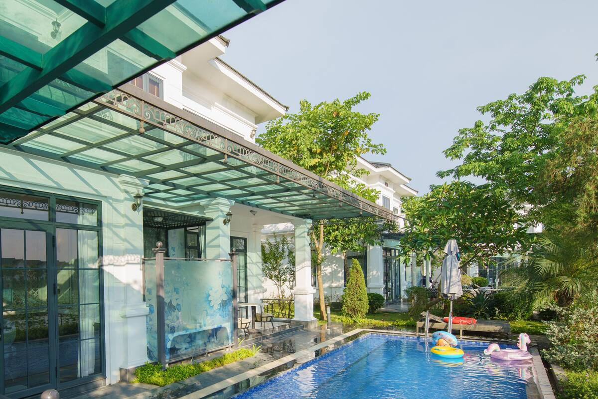 A swimming pool with a glass enclosure, surrounded by greenery and a building with a white facade.