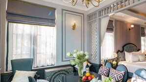 A hotel room with a bed, a dining table with fruit and a vase, a chair, and a decorative wall panel.