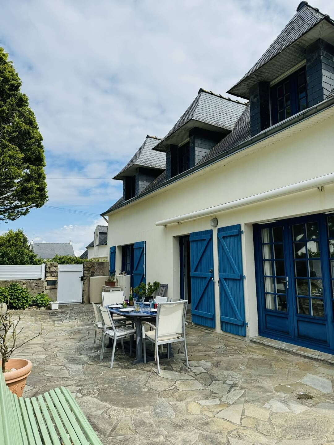 A two-story building with a stone patio, a dining table with chairs, and blue shutters on the windows.