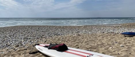A beach with a surfboard and wetsuit on the sand, a pebble beach, and the ocean in the background.
