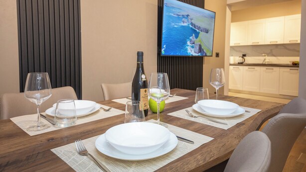 A dining table set with plates, glasses, and a bottle, with a TV mounted on the wall displaying a scenic view. - Blloku Unique Apartments 2 (Tirana)