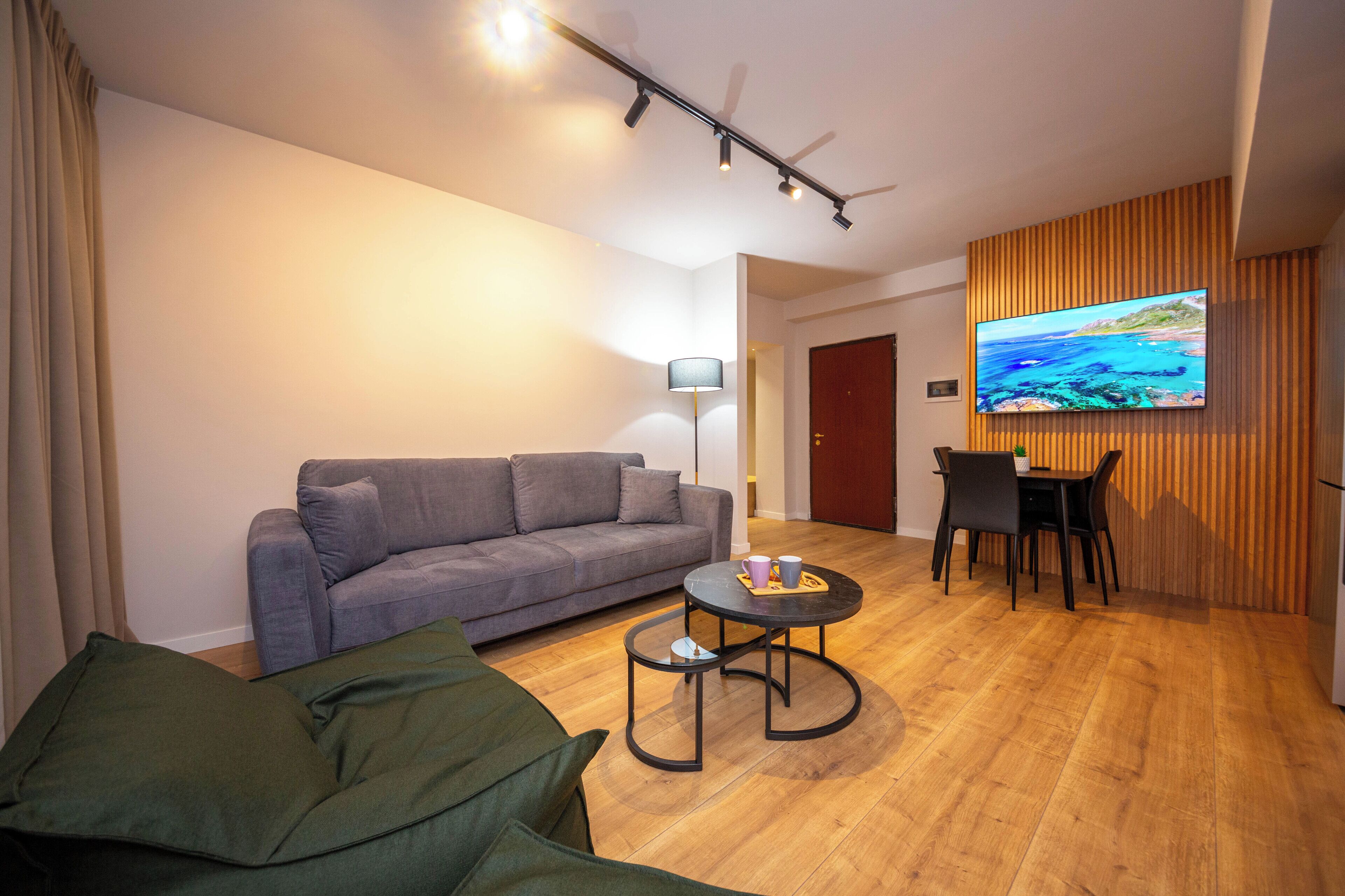 Una sala de estar moderna con un sofá gris, una mesita redonda con una bandeja, un televisor instalado en una pared de madera, y un comedor con sillas.
