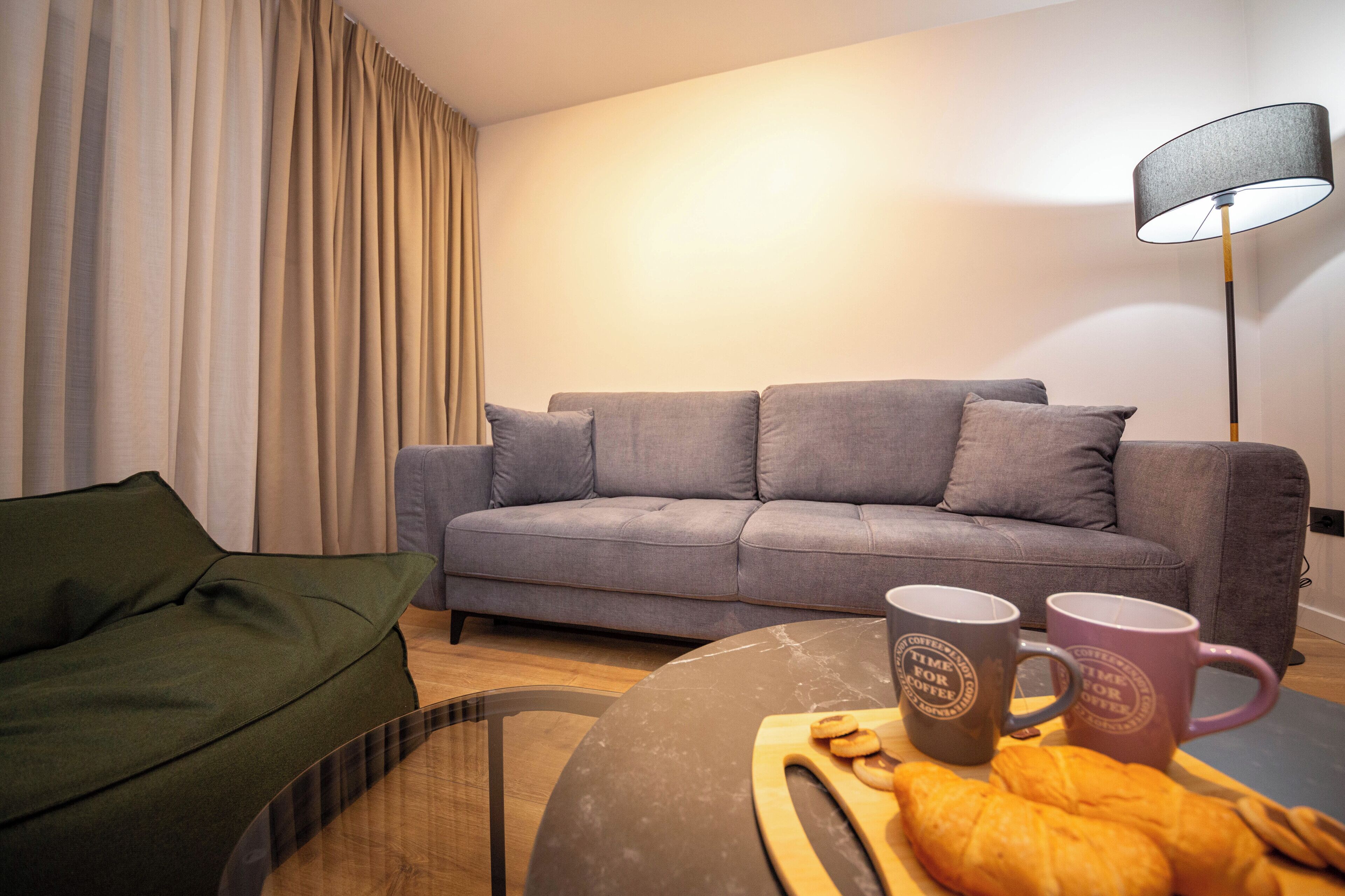 A living room with a grey sofa, a green armchair, a coffee table with a croissant and two mugs, and a floor lamp.