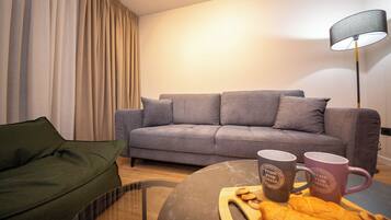 A living room with a grey sofa, a green armchair, a coffee table with a croissant and two mugs, and a floor lamp.