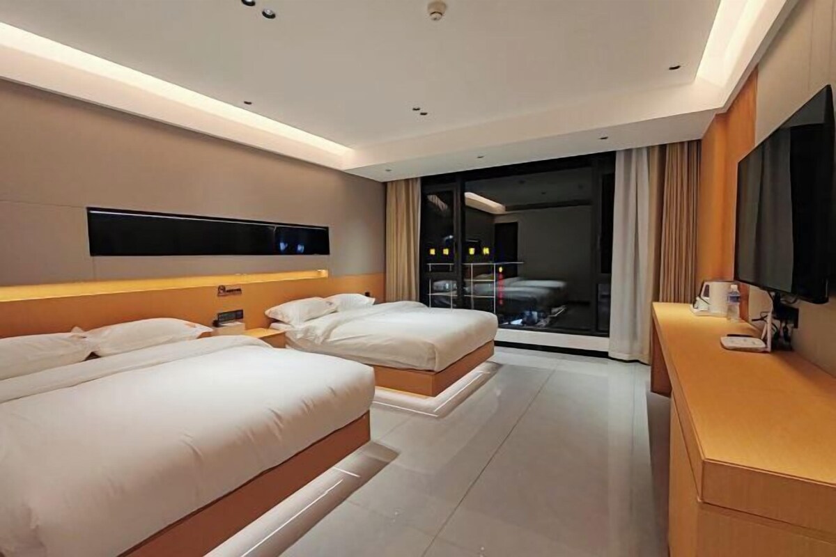 Una habitación de hotel moderna con dos camas, un televisor de pantalla plana montado en la pared, un escritorio con una lámpara y un ventanal con cortinas.